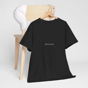 Puede incluir: Una camiseta negra con el texto blanco "ZERO EXCUSES". La camiseta está colgada en una percha de madera. Una gorra de béisbol blanca descansa sobre el respaldo de una silla de madera.