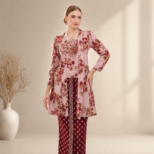 May include: A light pink long-sleeved tunic featuring a vibrant floral print in shades of red, orange, and green, paired with a dark maroon skirt with a white geometric pattern. The tunic has a layered front panel and is worn with a multi-strand pearl necklace.