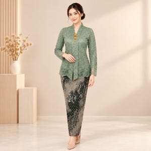 Peut inclure: Un chemisier à manches longues en dentelle vert sauge avec une encolure en V et une broche dorée au centre, associé à une jupe longue aux tons sombres présentant un motif floral et abstrait détaillé dans des nuances de noir, gris et marron.