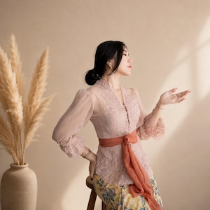 Puede incluir: Una blusa de encaje rosa empolvado de manga larga con cuello en V y mangas transparentes, que incluye un fajín de color naranja quemado atado a la cintura. Se combina con una falda amarilla estampada con un diseño floral en tonos azules y verdes.