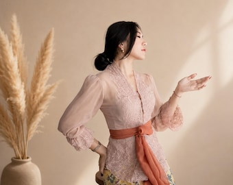 May include: A long-sleeved dusty pink lace blouse with a V-neckline and sheer sleeves, featuring a solid burnt orange sash tied at the waist. The blouse is paired with a yellow patterned skirt that displays a floral design in blue and green tones.