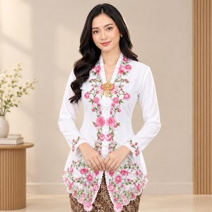 Puede incluir: Una blusa kebaya blanca de manga larga con intrincados bordados florales rosas a lo largo de la tapeta frontal y el dobladillo. La prenda incluye un borde de encaje festoneado, un escote en V y un broche dorado centrado en el pecho.