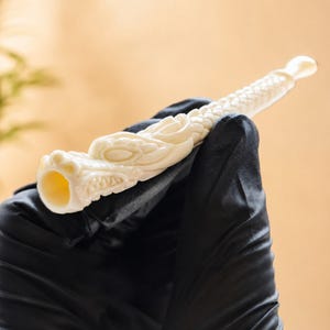 May include: An off-white, carved smoking pipe featuring a detailed dragon head at the bowl end. The stem of the pipe displays a textured, scale-like pattern that extends along the length of the body, ending in a smooth, rounded mouthpiece.