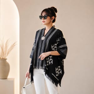 Puede incluir: Un poncho negro con rayas verticales blancas y un patrón geométrico de cuadrados. La prenda tiene un cuello en V con un broche plateado decorativo, mangas tres cuartos y un dobladillo con flecos. Se lleva sobre unos pantalones blancos.