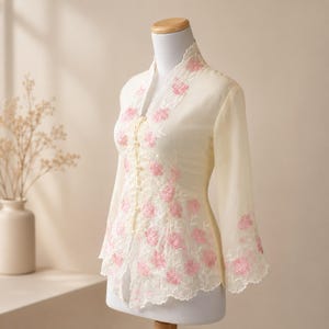 Puede incluir: Una blusa de manga larga color crema con cuello mao y cierre frontal de botones con presillas. La prenda está decorada con un detallado bordado floral rosa y patrones de hojas blancas, terminada con un dobladillo festoneado.