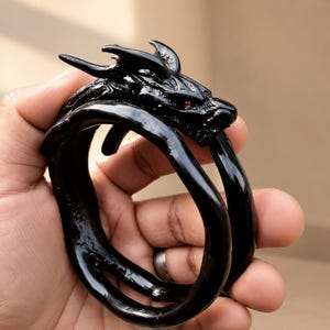 May include: A glossy black bracelet shaped like a coiled dragon with a textured head, curved horns, and small red eyes. The dragon's body forms the circular band of the bracelet, featuring a smooth, reflective finish throughout the entire piece.