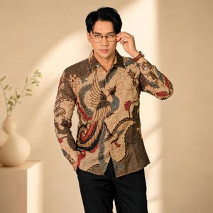 May include: A long-sleeved button-down shirt in a light tan hue, featuring a detailed batik-style print of a phoenix in shades of blue, red, and dark brown. The shirt has a classic collar, a front button placket, and fitted cuffs on the sleeves.