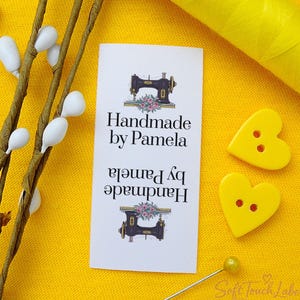 Può includere: Un'etichetta bianca con la scritta "Handmade by Pamela" e un'illustrazione di una macchina da cucire vintage. Due bottoni gialli a forma di cuore e una bobina di filo giallo su sfondo giallo. Sono visibili anche un rametto floreale e uno spillo.