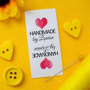 Può includere: Un'etichetta bianca con la scritta "HANDMADE by Louise" e un cuore rosa, circondata da accessori da cucito gialli. Include bottoni a forma di cuore, un bottone rotondo, uno spillo e una bobina di filo giallo, il tutto su sfondo giallo.