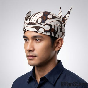 May include: A traditional Javanese blangkon headpiece featuring a brown and white batik pattern with intricate geometric and floral motifs. The headgear has a rounded crown and two pointed, upright fabric extensions at the back of the head.
