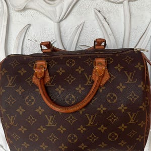 Könnte beinhalten: Braune Handtasche mit sich wiederholendem Monogrammmuster und hellbraunen Ledergriffen und -besatz. Die Tasche hat einen Reißverschluss und wird vor einem weißen Hintergrund mit floralem Relief gehalten.