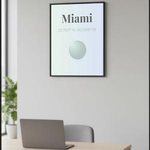 May include: A framed poster with the word "Miami" in bold, black text, along with coordinates. Below, a light green and blue gradient background features a white sphere. A laptop sits on a light wood desk with a gray chair.