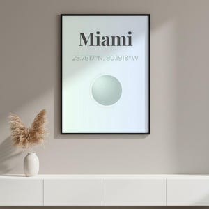 May include: A framed art print with the word "Miami" in a bold font, along with its coordinates. The print has a gradient background and a circular design element. A white vase with dried plants sits on a white cabinet below.