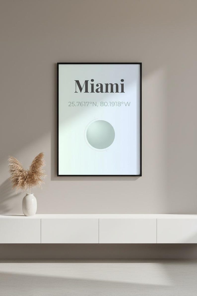 May include: A framed art print with the word "Miami" in a bold font, along with its coordinates. The print has a gradient background and a circular design element. A white vase with dried plants sits on a white cabinet below.