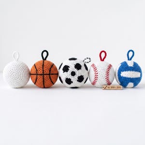 May include: A collection of five crocheted sports balls: a white ball, an orange basketball, a soccer ball, a baseball, and a blue volleyball. Each ball has a loop for hanging. The baseball has red stitching, and the soccer ball has black pentagons.