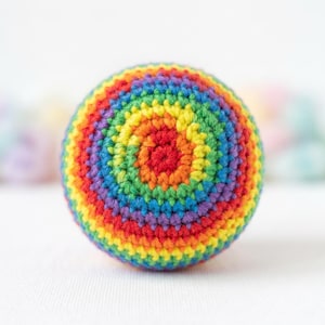 May include: A crocheted ball with a spiral pattern of vibrant rainbow colors. The ball features concentric circles of red, orange, yellow, green, blue, and purple. The handmade item is set against a soft, white background.