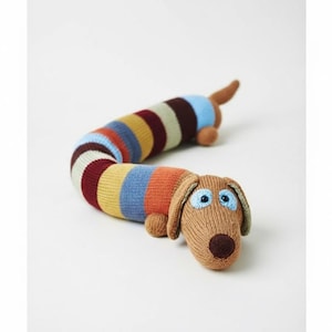 Peut inclure: Un jouet pour chien teckel tricoté, avec un corps rayé multicolore. La tête, les oreilles et la queue sont marron, avec des yeux bleus. Le jouet est conçu pour ressembler à un chien saucisse.