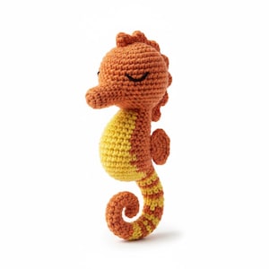 May include: A crocheted seahorse toy in orange and yellow. The seahorse has a textured design, closed eyes, a curved tail, and a rounded belly. The toy is designed to stand upright. The measurements are in centimetres.