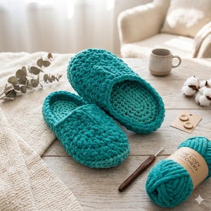 Puede incluir: Un par de pantuflas de ganchillo turquesa se encuentra sobre una superficie de madera, junto a una madeja de hilo turquesa y un ganchillo. Las pantuflas están hechas de un hilo grueso y texturizado. La etiqueta del hilo dice 'The Cozy Stitchery'.