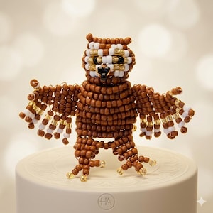May include: A beaded owl figurine with outstretched wings. The owl is primarily brown with white and gold accents on the wings and head. The eyes and beak are black. The owl is standing on a white, circular base.
