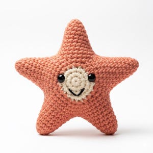 May include: A coral-colored, crocheted starfish toy with a smiling face. The starfish has five points and a cream-colored center with black eyes and a stitched black smile. The toy is made of a textured yarn and sits on a white background.