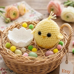 Puede incluir: Un pollito de ganchillo hecho a mano se encuentra en una cesta de mimbre con una cáscara de huevo de ganchillo blanca y pompones de colores. La cesta contiene paja artificial y huevos de Pascua decorativos. Una etiqueta de Etsy está adjunta.