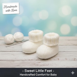 Op de afbeelding: Een paar handgebreide witte babyslofjes met een omgeslagen boord, gepresenteerd op een licht houten oppervlak. Twee bollen witte wol en een haaknaald liggen naast de slofjes. De tekst "Handmade with Love" is inbegrepen.