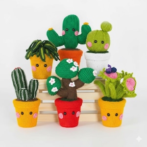 May include: A selection of hand-knitted cacti and other plants in various pots. The plants are green, yellow, and white, with pink cheeks and black button eyes. The pots are orange, yellow, red, and white, on a wooden crate.