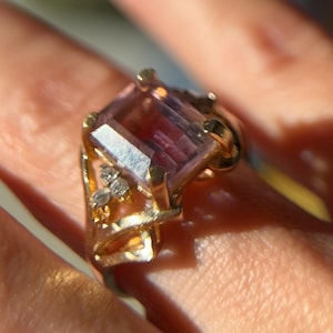 May include: A gold ring featuring a large, rectangular, faceted, pink gemstone. The ring has a gold band and is accented with small, clear gemstones on the side. The ring is shown close up, with the details of the setting and stone clearly visible.