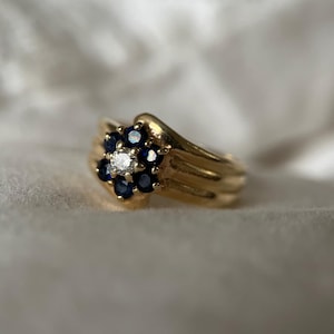 Puede incluir: Un anillo de oro con un diseño floral. La flor está compuesta por piedras preciosas de color azul oscuro que rodean una piedra preciosa central y transparente. El anillo tiene un acabado liso y pulido y una banda curva.