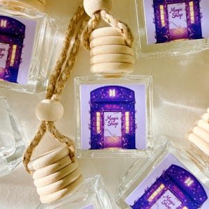 May include: Clear glass car air fresheners with a wooden top and a decorative image of a purple door with the text "Magic Shop". The air fresheners are hanging from a braided rope.