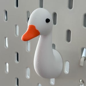 Op de afbeelding: Een witte, gansvormige wandhaak met een oranje snavel en zwarte ogen. De haak is gemonteerd op een witte pegboard achtergrond. De gans is een decoratief item.