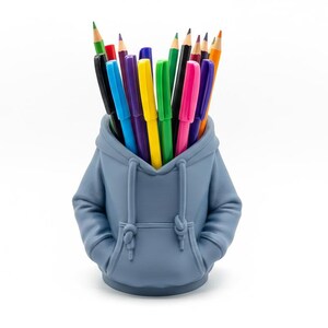 May include: A blue hoodie-shaped desk tidy filled with colourful pens and pencils. The organiser has a hood, pockets, and a drawstring. The pens and pencils are various colours, including red, green, yellow, and pink. The background is white.
