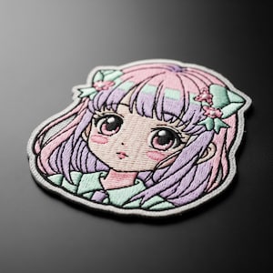 Puede incluir: Parche bordado con una chica de estilo anime con cabello rosa y morado, ojos grandes rosados y un lazo verde claro. El parche tiene un borde blanco y está sobre una superficie negra.
