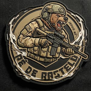 Könnte beinhalten: Ein kreisförmiger taktischer Aufnäher mit einem braunen Bären in beigefarbener Tarnuniform, Helm und Headset, der ein schwarzes Gewehr hält. Der Text JEDUIÉ-BR und PÉ DE RASTELO ist in Schwarz auf dem beigen Hintergrund gedruckt.
