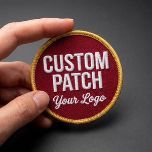 Puede incluir: Parche redondo personalizado con el texto "CUSTOM PATCH Your Logo" en blanco, bordado sobre un fondo granate. El parche tiene un borde dorado.