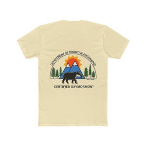 May include: A cream-colored t-shirt featuring a graphic of a black tapir standing before blue mountains, green pine trees, and an orange sun. The text reads DEPARTMENT OF COGNITIVE DISSONANCE, EST. 1830, and CERTIFIED OXYMORMON at the bottom.