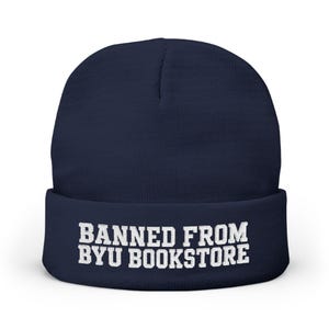 May include: A navy blue cuffed beanie featuring the white embroidered text BANNED FROM BYU BOOKSTORE centered on the front fold. The knit hat has a rounded crown and a thick, folded brim that displays the bold, capitalized lettering clearly.