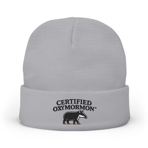 May include: A light gray cuffed beanie featuring black embroidered text that reads CERTIFIED OXYMORMON followed by a small graphic of a black tapir standing on a thin brown line, centered on the front of the folded cuff of the knit hat.
