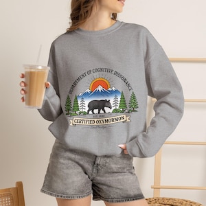 May include: A heather gray crewneck sweatshirt featuring a graphic print of a black bear standing in front of mountains and pine trees. The text reads Department of Cognitive Dissonance, Est. 1830, Certified Oxymormon, and Soul Nudge.