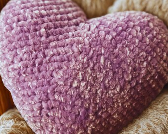 May include: A heart-shaped, lavender-colored pillow with a textured, plush surface. The pillow is resting on a cream-colored, knitted blanket. The pillow is a decorative item.