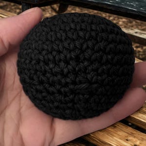 May include: A small, spherical object crafted from black yarn using a dense crochet stitch pattern. The item features a uniform, textured surface created by the interlocking loops of the dark thread, forming a compact and rounded shape.