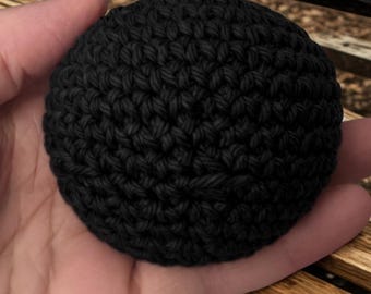 May include: A small, spherical object crafted from black yarn using a dense crochet stitch pattern. The item features a uniform, textured surface created by the interlocking loops of the dark thread, forming a compact and rounded shape.