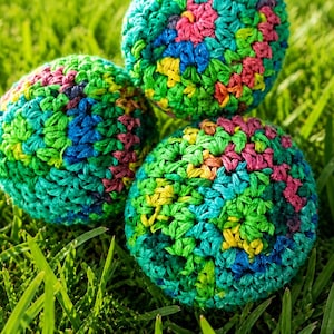 May include: Three colorful crocheted balls rest on a bed of green grass. The balls are a mix of green, blue, pink, and yellow yarn, creating a textured, handmade appearance. The balls are round and appear to be decorative or playful.