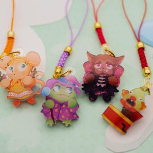 May include: Four acrylic charms with colourful braided phone straps. The charms feature a yellow mouse-like character, a green frog in a purple hat, a brown beast-like figure with pink wings, and a clown popping out of a red jack-in-the-box.