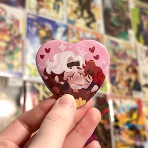 May include: A heart-shaped pin featuring a colorful illustration of two characters with brown and white hair embracing. The background is light pink with small red hearts and white star accents. The characters have flushed cheeks and closed eyes.