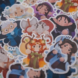 May include: A collection of die-cut vinyl stickers featuring various chibi-style fantasy characters. The designs include figures with diverse hair colors, outfits, and accessories like axes, staves, and glasses, all rendered in a soft, colorful art style.