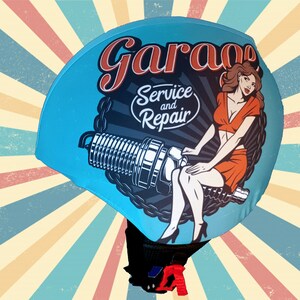 May include: A blue helmet with a vintage-style graphic of a woman sitting on a spark plug. The graphic includes the text "Garage Service and Repair".