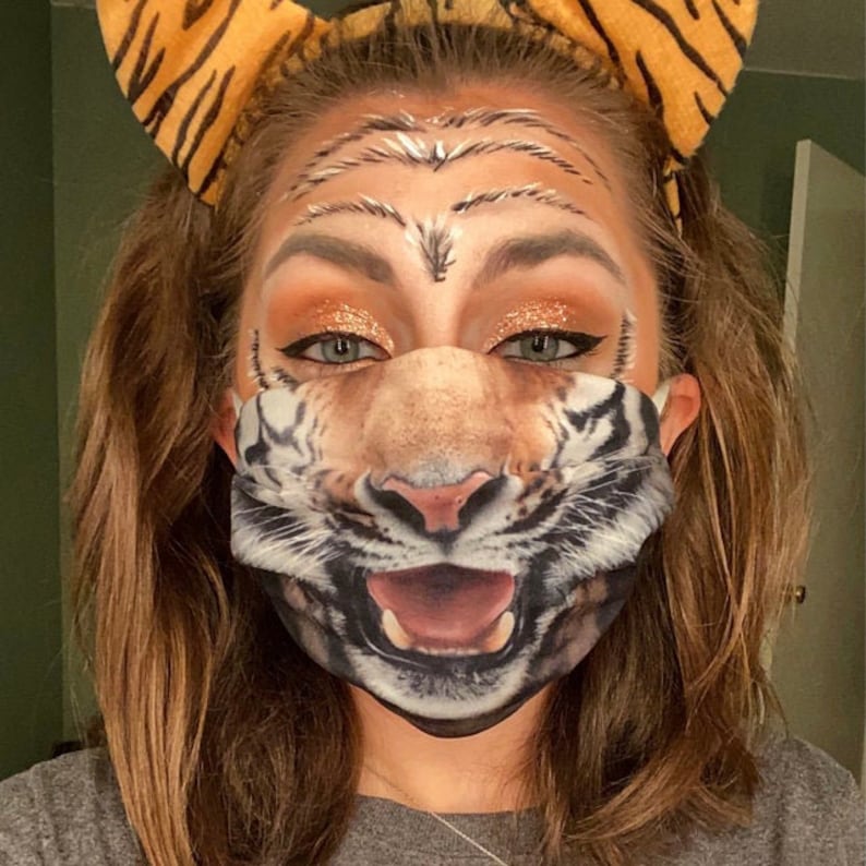 May include: A woman wearing a tiger-print face mask with a matching headband. Her face is painted with tiger stripes and glitter makeup.