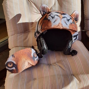 May include: A brown and black tiger print helmet with a matching eye mask. The helmet has a black adjustable strap and a black foam lining. The eye mask is made of soft fabric and has a tiger print design.